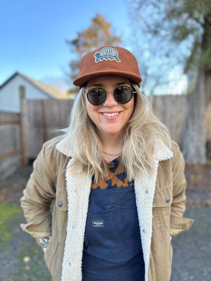 The Athletic Armadillo Hat in rust features an unstructured 6-panel design with an adjustable fabric strap and brass metal clasp. Embroidered with a stylized armadillo logo, this hat combines comfort and style for everyday wear