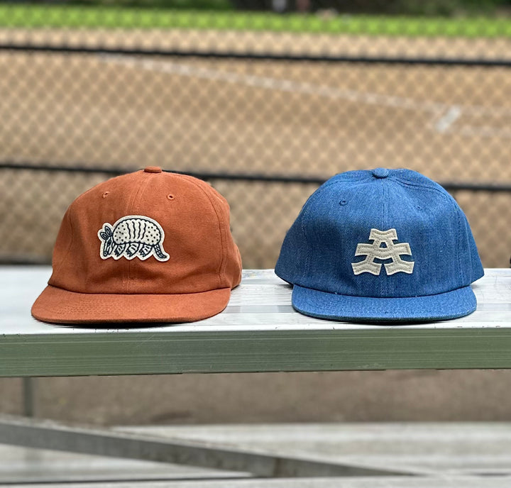 The Athletic Armadillo Logo Hat in rust features an unstructured 6-panel design with an adjustable fabric strap and brass metal clasp. Embroidered with a stylized armadillo logo, this hat combines comfort and style for everyday wear