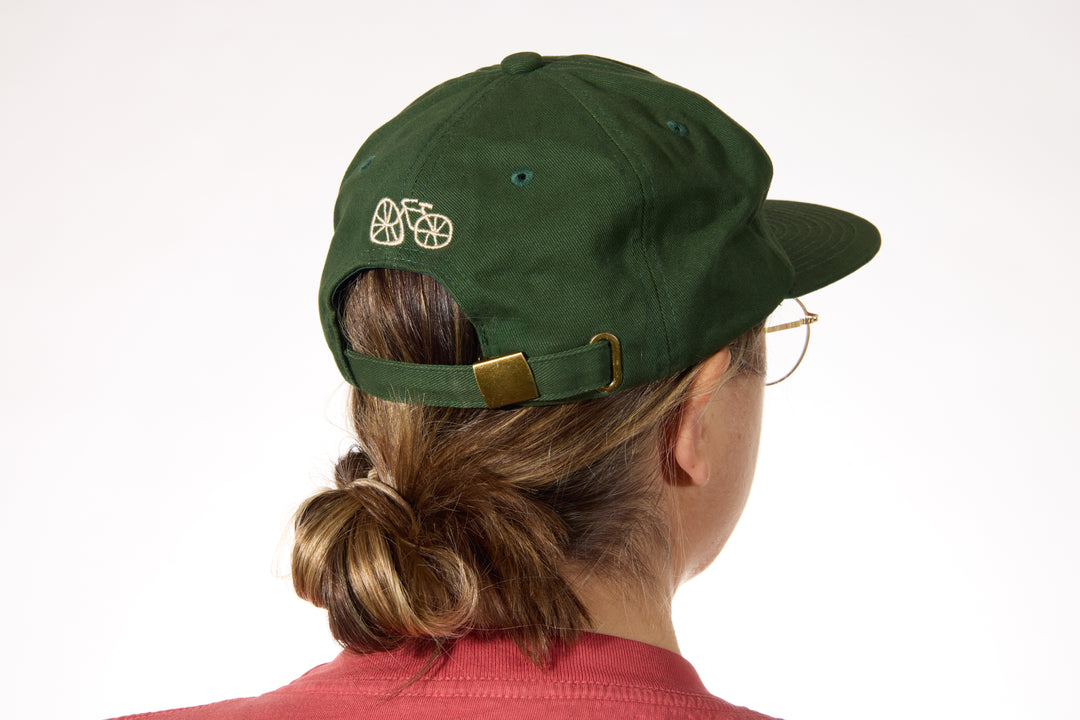 The I LIKE BIKE Hat is a green, 6-panel cap featuring the embroidered phrase "I LIKE BIKE" in white. It includes an adjustable fabric strap with a brass metal clasp for a customizable fit. Made from 100% cotton, this hat offers comfort and style for cycling enthusiasts