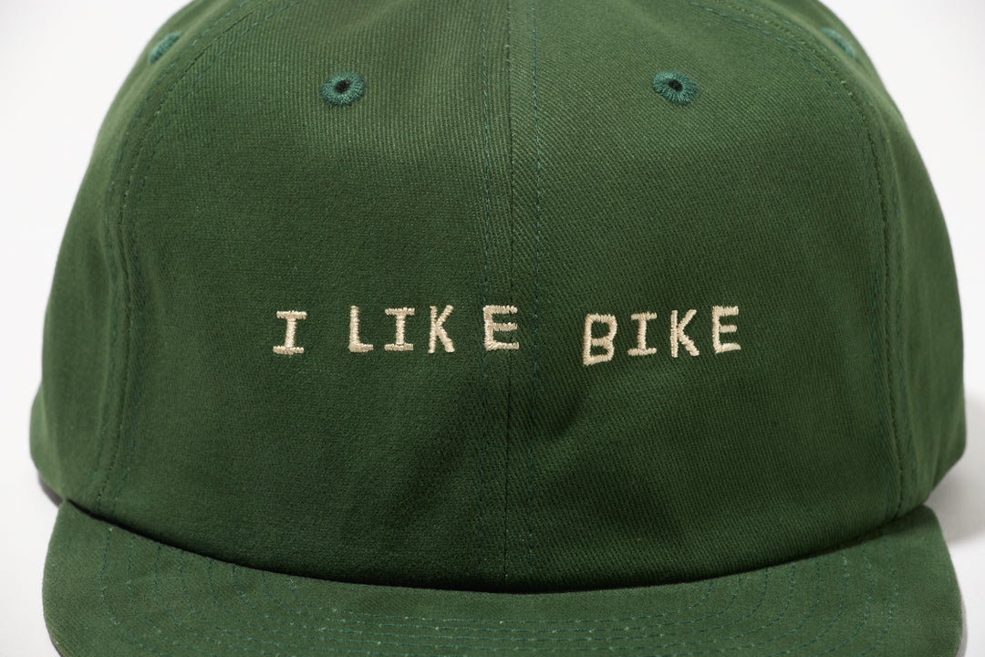 The I LIKE BIKE Hat is a green, 6-panel cap featuring the embroidered phrase "I LIKE BIKE" in white. It includes an adjustable fabric strap with a brass metal clasp for a customizable fit. Made from 100% cotton, this hat offers comfort and style for cycling enthusiasts