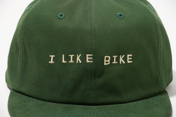 The I LIKE BIKE Hat is a green, 6-panel cap featuring the embroidered phrase "I LIKE BIKE" in white. It includes an adjustable fabric strap with a brass metal clasp for a customizable fit. Made from 100% cotton, this hat offers comfort and style for cycling enthusiasts