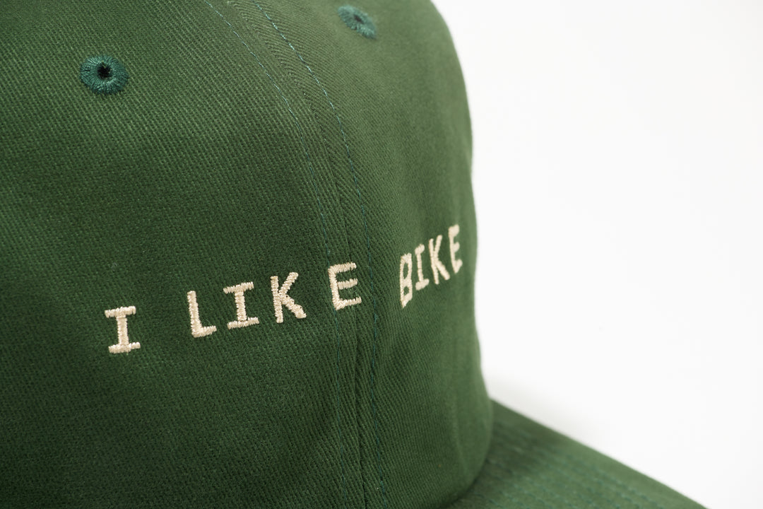 The I LIKE BIKE Hat is a green, 6-panel cap featuring the embroidered phrase "I LIKE BIKE" in white. It includes an adjustable fabric strap with a brass metal clasp for a customizable fit. Made from 100% cotton, this hat offers comfort and style for cycling enthusiasts