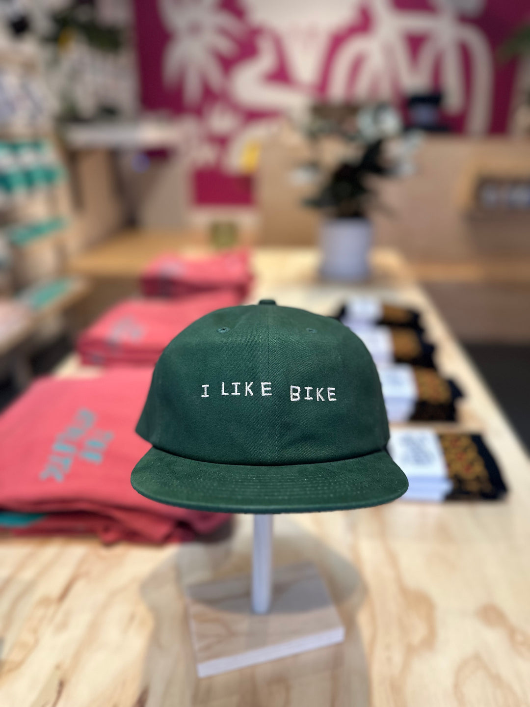The I LIKE BIKE Hat is a green, 6-panel cap featuring the embroidered phrase "I LIKE BIKE" in white. It includes an adjustable fabric strap with a brass metal clasp for a customizable fit. Made from 100% cotton.