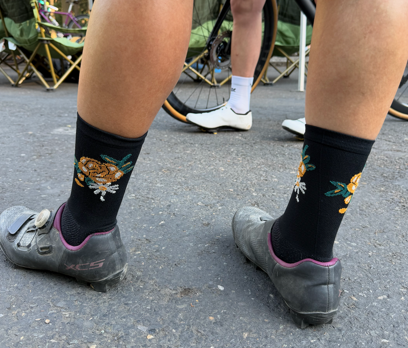 Black cycling socks with embroidered floral  patterns worn with gray cycling shoes on pavement.
