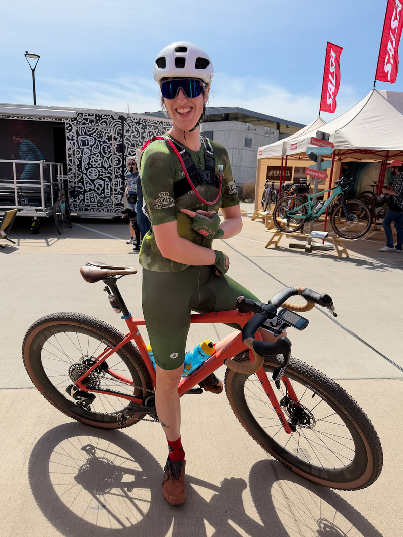 A smiling woman wearing a white helmet, sunglasses, and an olive green cycling jersey and shorts stands outdoors next to an orange mountain bike. She has a hydration pack on her back and is posing in a sunny outdoor event or shop setting with tents, bikes, and a trailer in the background.