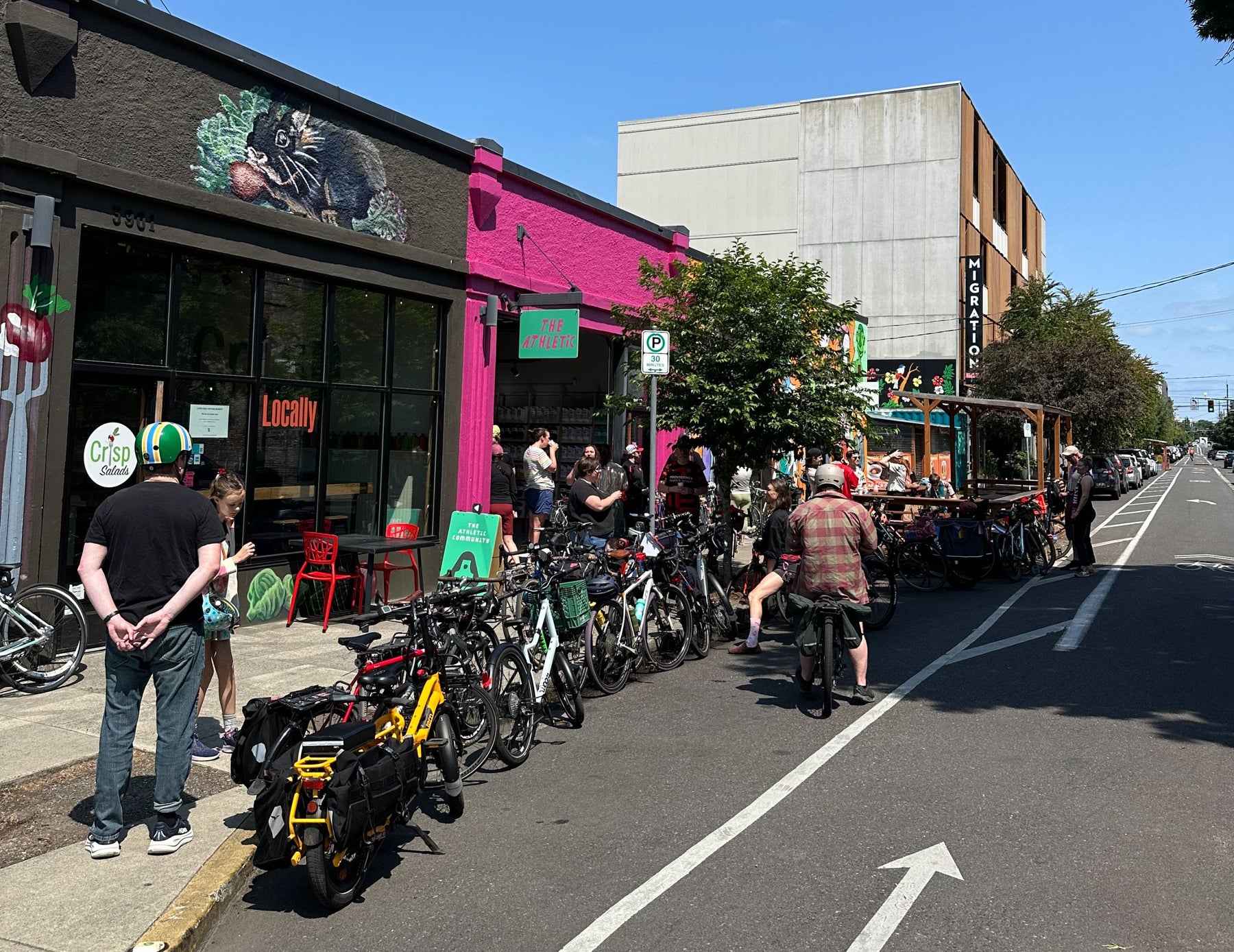 People standing on a sidewalk outside The Athletic with bicycles parked along the street in front of a building with a pink facade.
