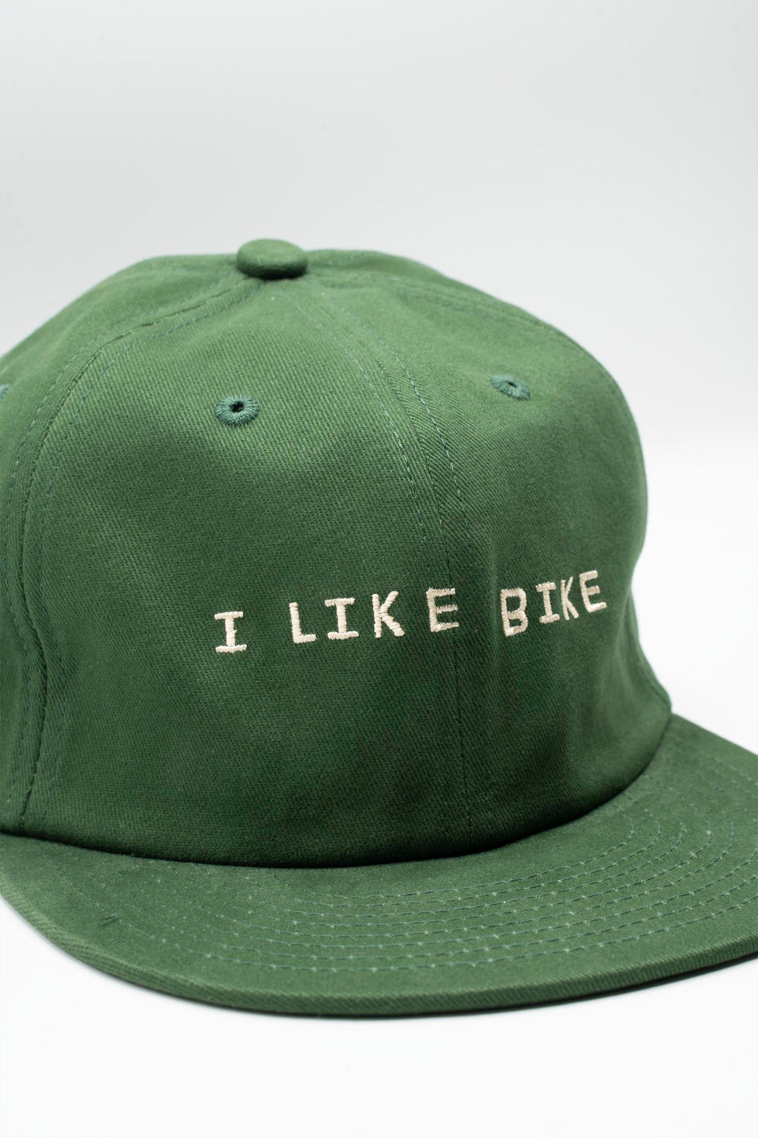 The I LIKE BIKE Hat is a green, 6-panel cap featuring the embroidered phrase "I LIKE BIKE" in white. It includes an adjustable fabric strap with a brass metal clasp for a customizable fit. Made from 100% cotton, this hat offers comfort and style for cycling enthusiasts