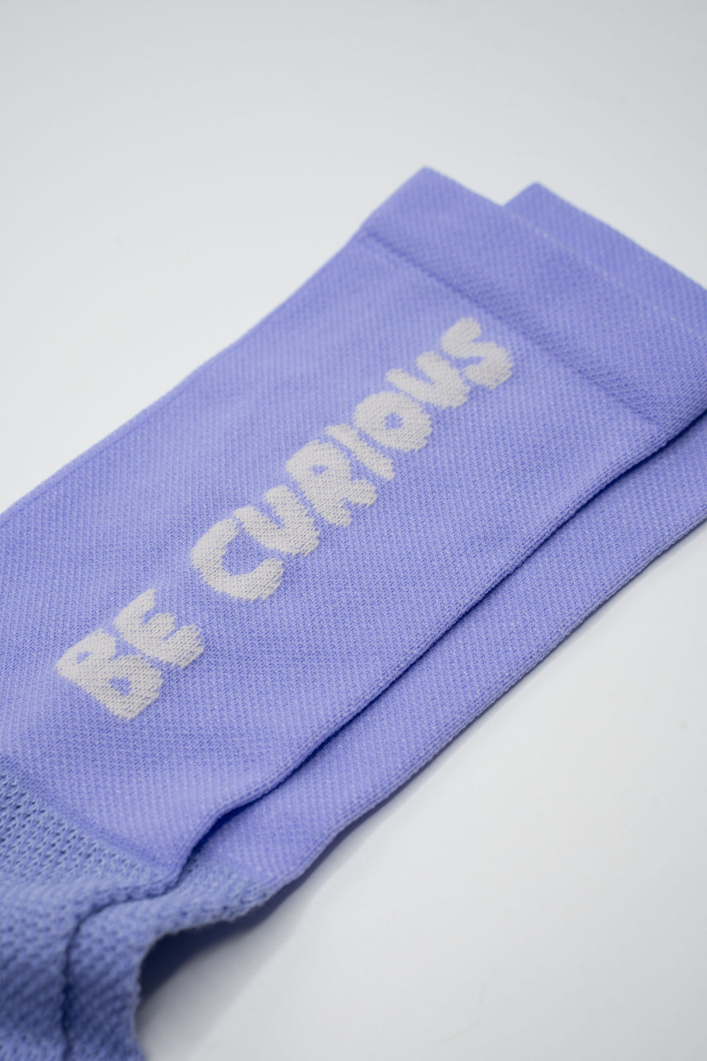 The Be Curious Socks are lavender socks featuring the phrase "Be Curious" on either side, designed for running and cycling. Perfect for cross-training, these socks combine style and performance. Please note that this item is on final sale and is not eligible for returns or exchanges